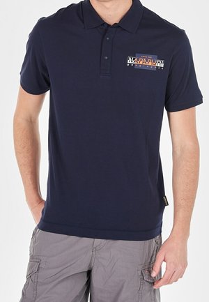 Mann trägt ein marineblaues Napapijri-Poloshirt mit kurzen Ärmeln und hellgraue Cargoshorts, steht mit einer Hand in der Tasche.
