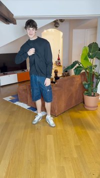 Pikad varrukad must särk, teksariidest cargo lühikesed püksid, hõbedased tossud. Siseruum, kus on puidust põrand ja pruunist korduurist diivan. Lähedal roheline taim.