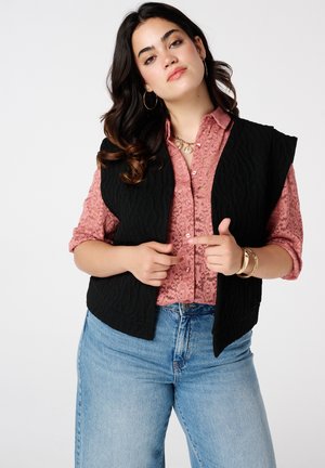 Zwarte gewatteerde vest gedragen over een gestructureerde roze blouse. Kenmerkt zich door een relaxte pasvorm, open voorkant en brede armsgaten. Het model draagt ook denim jeans.