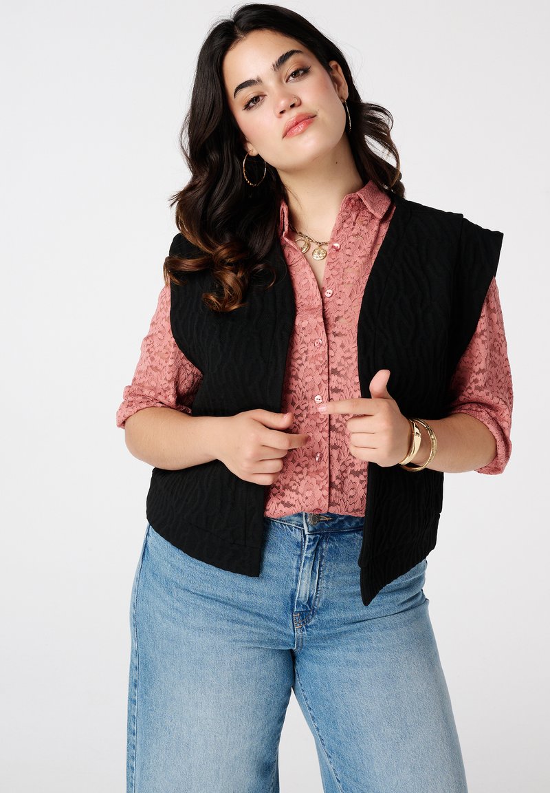 Zwarte gewatteerde vest gedragen over een gestructureerde roze blouse. Kenmerkt zich door een relaxte pasvorm, open voorkant en brede armsgaten. Het model draagt ook denim jeans.