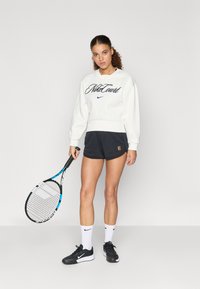 Λευκή μπλούζα με το λογότυπο "NikeCourt", συνδυασμένη με μαύρες σορτς. Το μοντέλο κρατάει μια ρακέτα τένις, φορώντας μαύρα αθλητικά παπούτσια και λευκές κάλτσες.