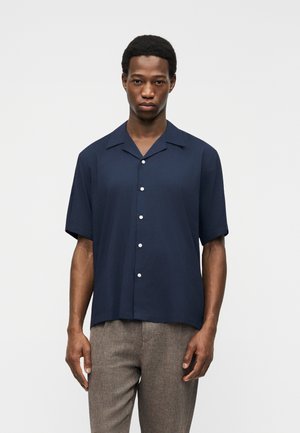 Jeune homme portant une chemise bleu marine à manches courtes boutonnée et un pantalon marron clair, debout devant un fond blanc uni.