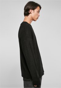 Suéter negro de punto oversize con textura acanalada y hombros caídos. La vista lateral muestra un ajuste relajado y mangas largas.