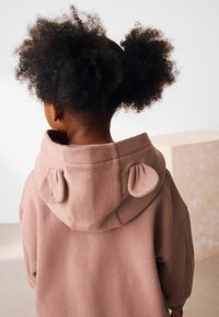 Haut à capuche en polaire rose avec une texture douce, doté d'une grande capuche ornée de deux accents arrondis ressemblant à des oreilles.