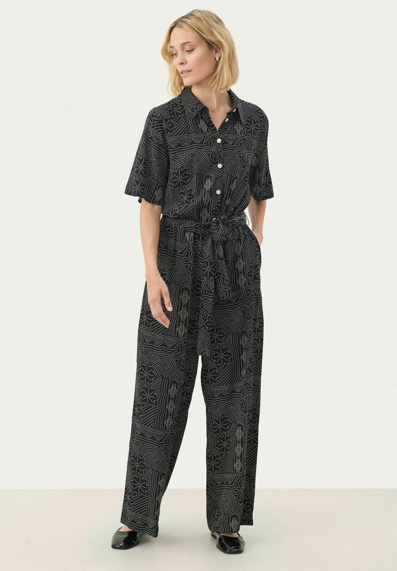 Black jumpsuit with short sleeves, button front, belt at the waist, and white geometric patterns throughout. Material appears lightweight and flowy.