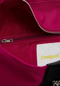 Intérieur de sac fuchsia avec une fermeture éclair argentée et une étiquette blanche comportant la marque "Desigual" en jaune cousue à l'intérieur.