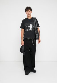 T-shirt preta de algodão com uma impressão gráfica, combinada com calças largas pretas, um cinto com studs e uma bolsa transversal preta.