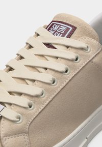 Κοντινό πλάνο μπεζ σουέντ αθλητικού παπουτσιού με εκρού κορδόνια, μεταλλικά ροζέτα και καφέ λογότυπο στη γλώσσα.