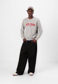 Szürke pulóver piros "Pepe Jeans" logóval, fekete széles nadrággal és bézs sportcipővel. A khaki sapka tökéletes kiegészítője a laza megjelenésnek.