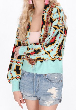 Mujer que lleva un cárdigan con flecos y estampado en azul claro, rojo, negro y amarillo sobre una camiseta rosa pálido con shorts de mezclilla desgastados.