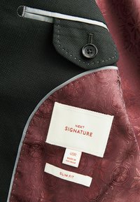 Tweeddul cu textură de culoare neagră, căptușeală din satin burgund cu un model embosat. Etichetele indică brandul, mărimea și detalii despre croi.