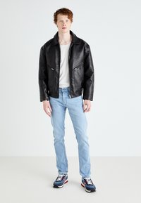 Casaco de couro preto com dois zíperes na frente, usado sobre uma camisa branca, combinado com jeans azul claro e ténis multicoloridos.