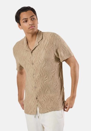 Młody mężczyzna ubrany w beżową teksturowaną koszulę na krótki rękaw z guzikiem oraz białe spodnie, patrzący w lewo na jednolitym tle.