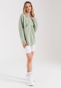 Jasnozielony, oversize'owy sweter z unikalnym wzorem wycięć, w połączeniu z białymi szortami rowerowymi z prążkowaniem i białymi trampkami.