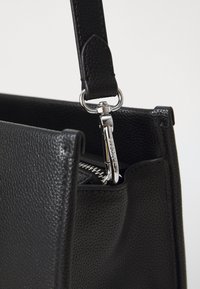 Sac en cuir noir texturé avec un clip en métal argenté et des détails de fermeture éclair. La sangle est réglable et cousue avec précision.