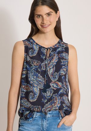 Femme aux longs cheveux foncés sourit, portant un haut sans manches bleu marine à motif cachemire avec liens à pompons et un jean bleu, debout devant un fond uni.