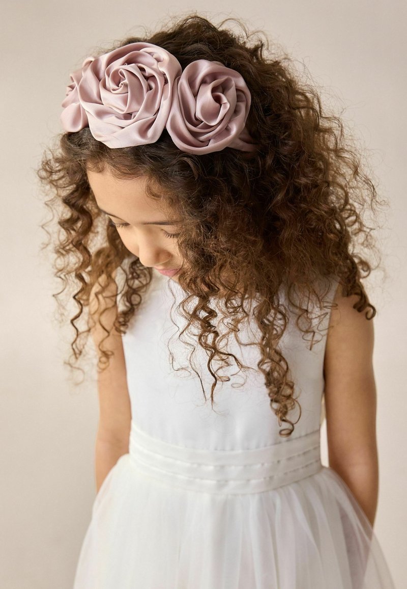 Un bandeau en satin rose orné de trois embellissements en forme de rose, porté par un enfant aux cheveux bouclés, sur un fond neutre.