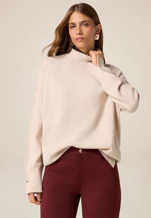 Lichtbeige oversized trui met een hoge ribbelboord, voorzien van een zachte textuur en zilveren knoopaccenten op de mouwen. Gecombineerd met bordeauxrode broek.