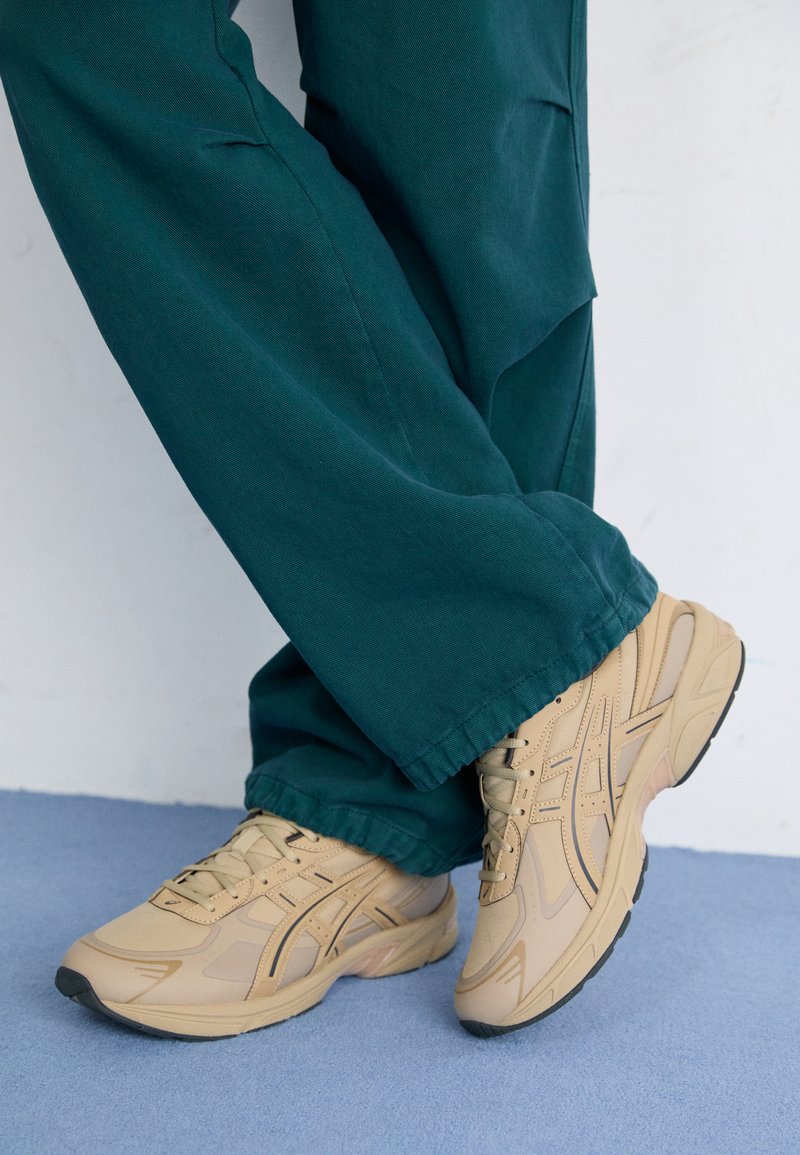Baskets de sport beiges avec une surface texturée, dotées de panneaux en maille, de semelles en caoutchouc légères et d'accents noirs, associées à un pantalon vert.