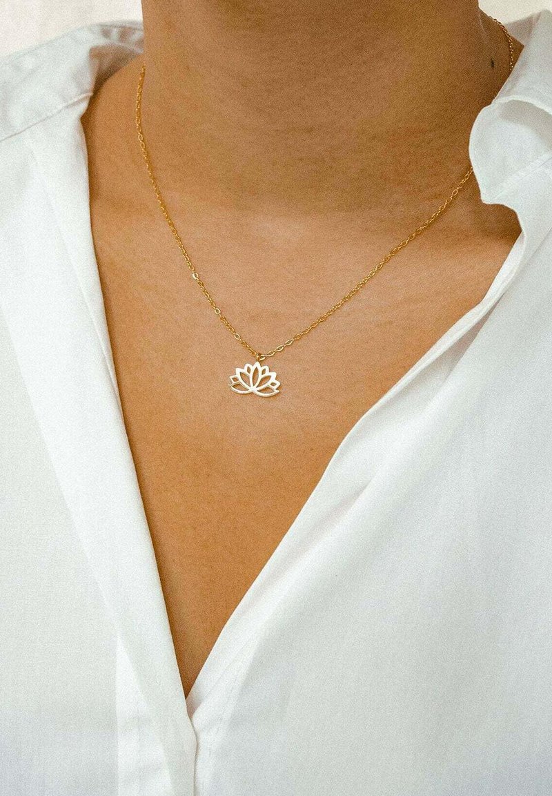 Collier en chaîne dorée avec un pendentif en forme de fleur de lotus présentant des détails découpés complexes, mis en valeur sur l'encolure d'une chemise blanche à col.