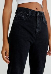 Gros plan d'une personne portant un jean noir taille haute et un haut court blanc, les mains détendues le long du corps et le ventre découvert.