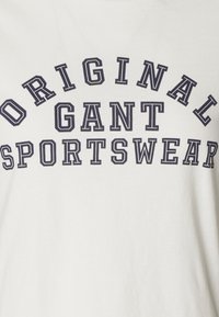 Bílý bavlněný tričko s výrazným námořnickým textem, který zní "ORIGINAL GANT SPORTSWEAR" v zakřiveném uspořádání přes hruď.