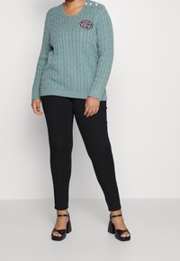 Ljus turkos stickad tröja med flätmönster, med tre vita knappar på vänster axel och en mörk broderad logotyp. Svarta smala byxor.