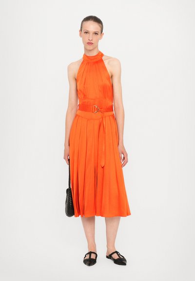 Oranžové šaty s halter krkom a skladmi, opásané v páse okrúhlou sponou. Modelka ich skombinovala s čiernou pletenou taškou a plochými topánkami.