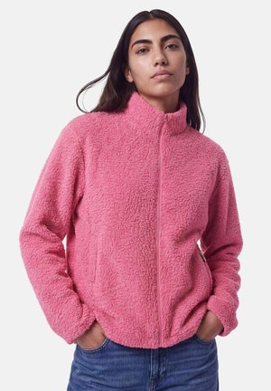 Mujer joven con cabello largo y oscuro, que lleva una chaqueta de forro polar texturizada de color rosa brillante y vaqueros azules, con las manos en los bolsillos, expresión neutral.