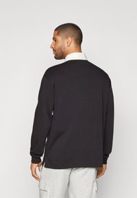 Črna majica z dolgimi rokavi in belim ovratnikom, izdelana iz mešanice mehkega bombaža. Ima sproščen kroj in rebraste manšete, prikazana na modelu s hrbtne strani.