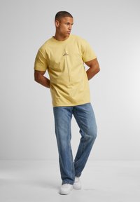 Camiseta de algodón amarilla con un diseño gráfico central en negro. Combinada con jeans de mezclilla azules y zapatillas blancas. Corte simple y relajado.
