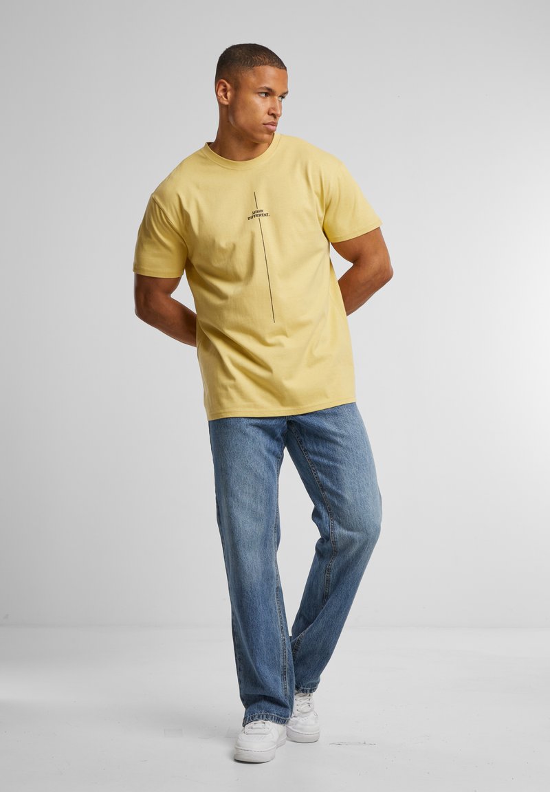 Yellow cotton t-shirt featuring a central black graphic text design. Paired with blue denim jeans and white sneakers. Simple relaxed fit.
