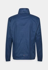 Chaqueta cortavientos azul marino hecha de un material ligero y brillante. Presenta un cuello alto y puños elásticos. Diseño minimalista.