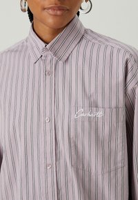 Light purple striped shirt with black vertical lines, buttoned collar, and a left chest pocket featuring white embroidered logo. Smooth fabric texture.