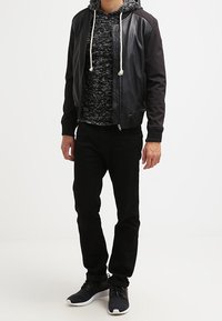Giacca bomber nera con maniche in pelle, felpa grigia con texture sotto, jeans neri e sneakers nere con suola bianca.