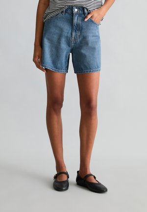 Person wearing mid-blue denim shorts, striped shirt, and black Mary Jane flats, standing against plain light gray background.