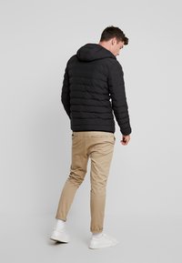 Chaqueta de plumas negra con capucha, con secciones acolchadas horizontales. Llevada sobre pantalones beige, con zapatillas blancas. Diseño sencillo.