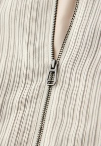 Fermeture éclair en métal partiellement dézippée sur un tissu beige avec de fines rayures verticales noires, tirette de la fermeture éclair gravée "STREET ONE".