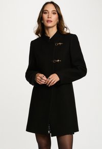 Abrigo negro con un cuello alto, dos cierres tipo toggles dorados, mangas largas y un corte recto. La tela tiene una textura suave, diseño minimalista.