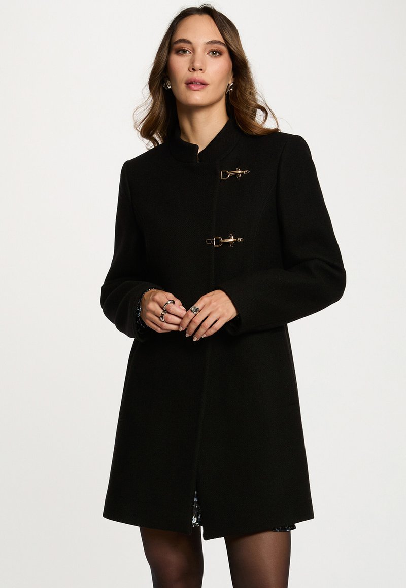 Abrigo negro con un cuello alto, dos cierres tipo toggles dorados, mangas largas y un corte recto. La tela tiene una textura suave, diseño minimalista.