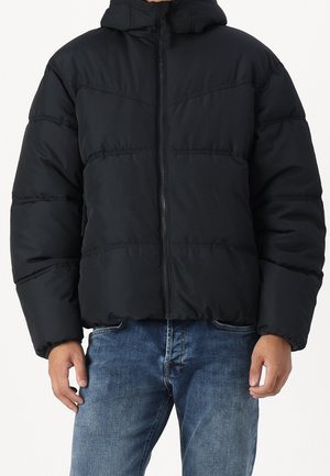 Mężczyzna w czarnej puchowej kurtce z przednim zamkiem błyskawicznym i niebieskich dżinsach na jednolitym tle.