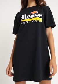 Schwarzes T-Shirt-Kleid, lockere Passform, kurze Ärmel. Mit auffälligem, in Weiß, Gelb und Rot gedrucktem Logo. Baumwollmaterial, glatte Textur.