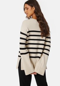 Beiger gestreifter Pullover mit schwarzen horizontalen Linien, langen Ärmeln, gerippten Bündchen und seitlichen Schlitzen. Weiches Material, lockere Passform und lässiges Design.