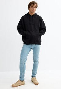 Sudadera con capucha negra con bolsillo canguro y cordón, combinada con vaqueros pitillo azul claro y zapatillas beige. Atuendo casual y cómodo.