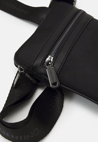 Borsa a tracolla nera in nylon con due zip, finitura testurizzata e tracolla con brand. Presenta hardware argentato e forma rettangolare slim.