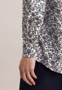 Koszula w kwiatowy wzór z długimi rękawami. Posiada białą bazę, misterny, wielobarwny kwiatowy wzór oraz mankiety na guziki. Materiał wygląda na gładki.