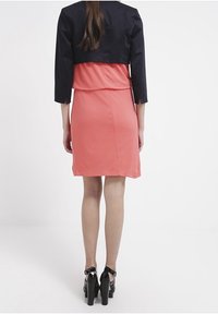 Korallklänning med en figursydd design, i kombination med en svart kort jacka. Svarta högklackade skor. Slät tygkvalitet, enkel skärning och trekvartsärmar.