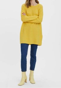 Żółty sweter z dzianiny z długimi rękawami i luźnym krojem, zestawiony z ciemnoniebieskimi dżinsami i jasnżółtymi botkami na obcasie.