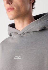Grå hettegenser laget av teksturert stoff, med en stor hette og en liten hvit "HUGO" logo på brystet. Nærbilde av halslinjen og stoffet.