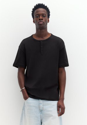 Jeune homme aux cheveux courts et bouclés, portant un t-shirt henley noir à manches courtes et un jean bleu clair, posant devant un fond blanc uni.